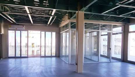 Open-plan office interior with green cellular steel beams, a glass-walled room, and sliding doors to a balcony.