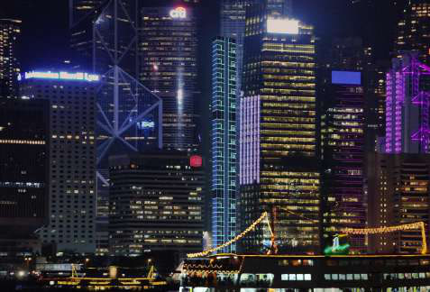 Hong Kong night skyline with a slender tower marked by a glowing blue grid, seen across the harbour.
