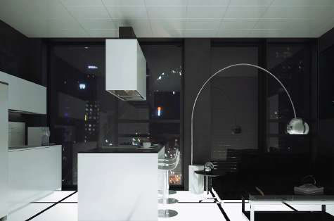 Black-and-white night interior: white kitchen island with stools, an arc lamp over a dark sofa, and city lights through full-height windows.