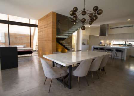 Interior of San Javier 245: dining table, rammed-earth wall, marble stair with glass railing, and kitchen beyond.