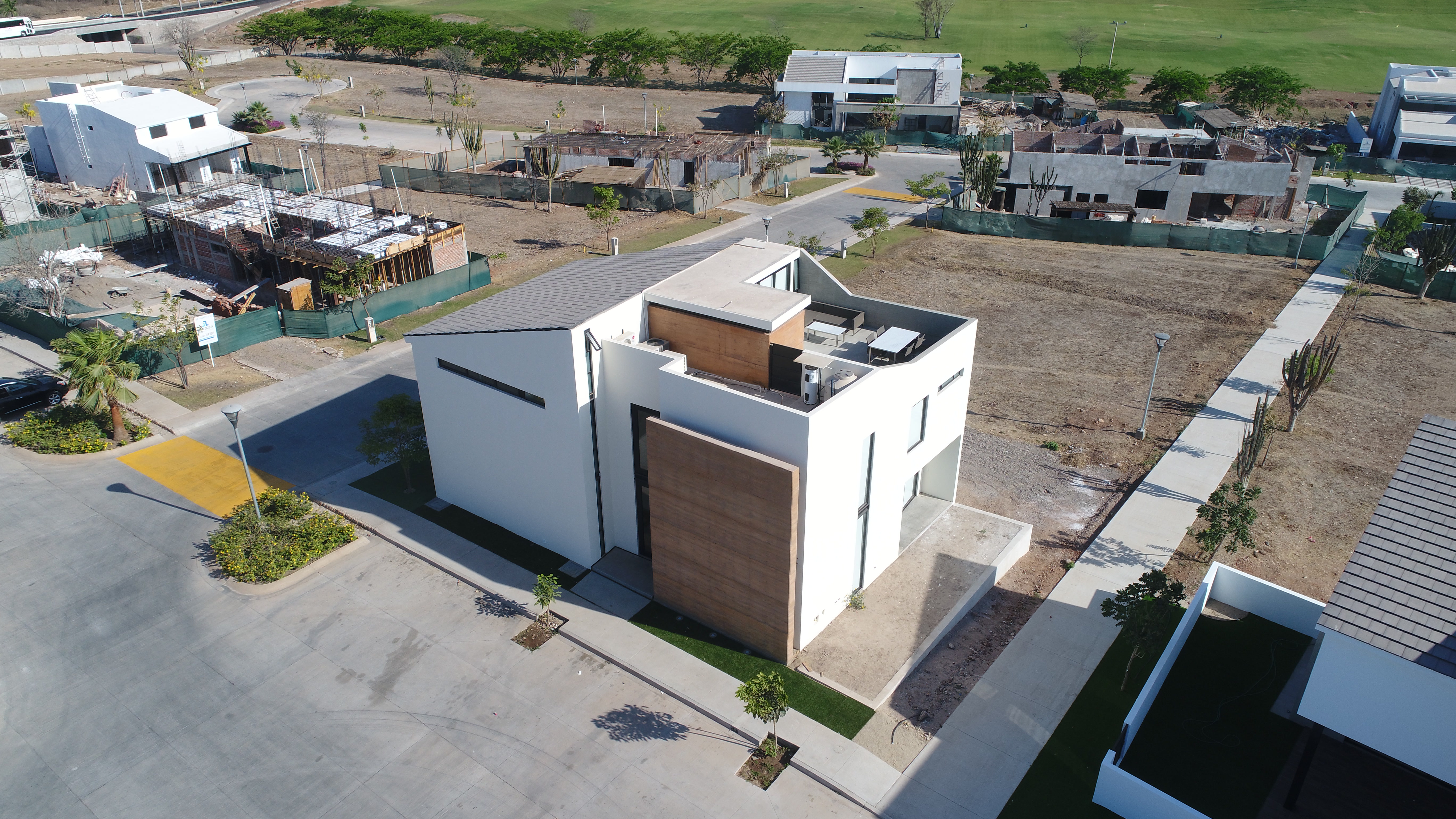 Aerial view of the white two-story house with a rammed-earth panel, rooftop terrace, and a neighbouring lot under construction.