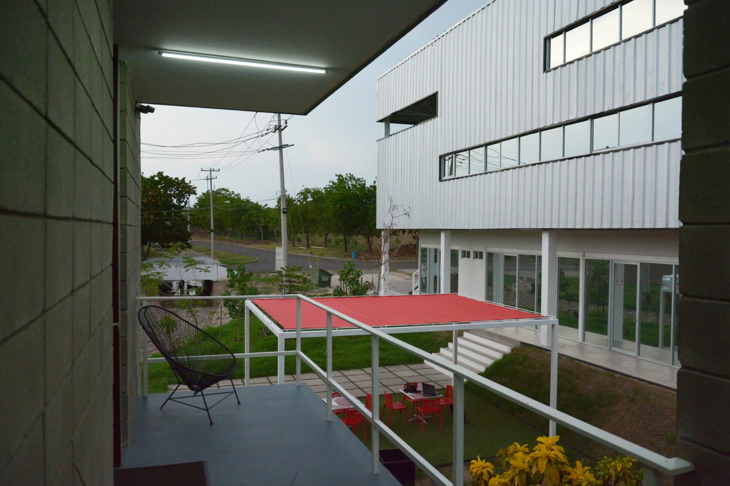View from a balcony across TARA’s courtyard: red awning, lawn, and a corrugated-metal volume with ribbon windows.