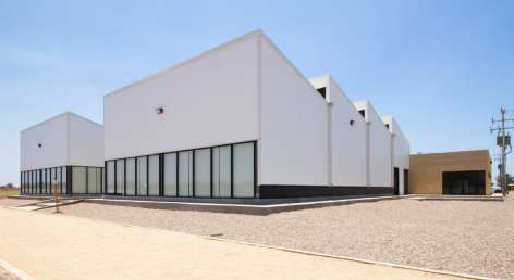 Viva Organica: white industrial volumes with a sawtooth roof and a glazed ground floor on a gravel site.
