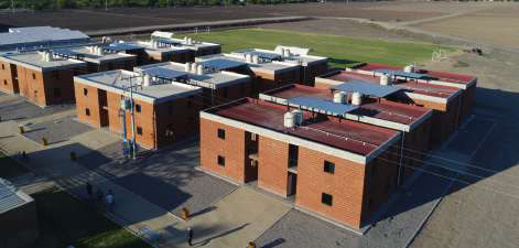 Aerial view of two-story brick housing blocks with rooftop tanks, walkways, and agricultural fields beyond.