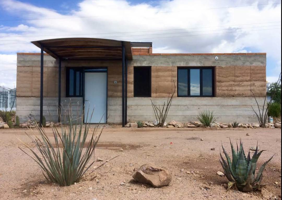 Single-story house with layered rammed-earth walls, a dark metal porch, and desert planting.